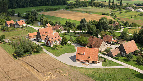 Fotoaufnahme eines Luftbildes der Baugruppe Ost. Die Häuser stehen in kleinen Gruppen und sind umgeben von Feldern, Wiesen und Bäumen. Im Hintergrund ist die weite Landschaft des Museums und der Umgebung erkennbar. 