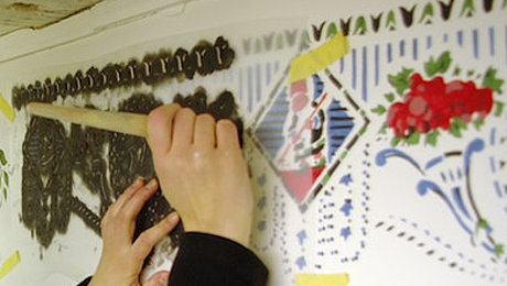 Fotoaufnahme einer Schablone, die mit Klebeband an einer Wand befestigt wurde. Das Muster der Schablone wird dabei mit Farbe und Pinsel stupfend auf die Wand gebracht. An der Wand befinden sich bereits Muster in verschiedenen Farben. 