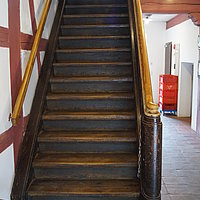 Fotoaufnahme des Treppenaufgangs im Gasthaus zur Krone aus Oberampfrach. Die 17-stufige Holztreppe mit dem Geländer auf beiden Seiten führt in das Obergeschoss. Am Fuß der Treppe steht ein Stuhl aus Holz, die Wände und die Decke sind im Fachwerkstil gehalten.