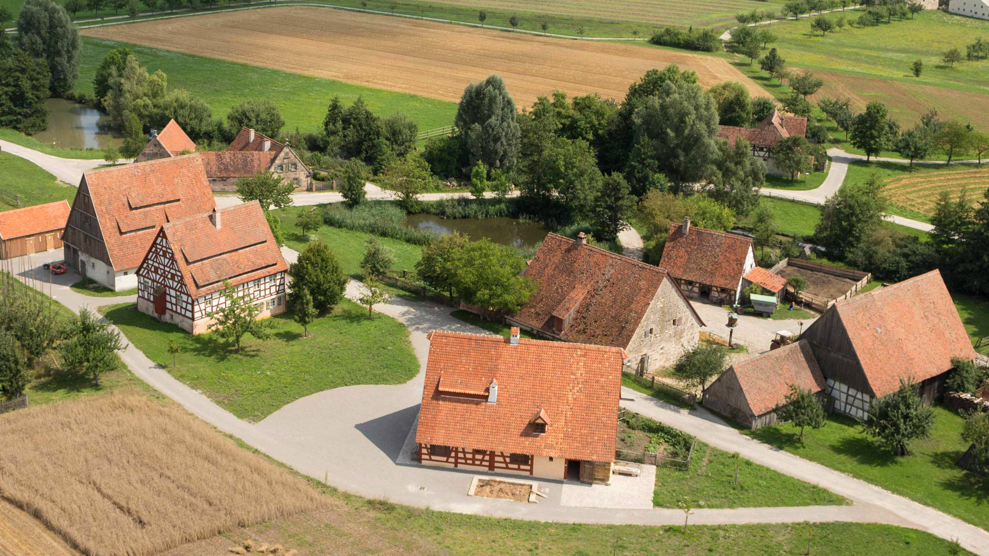 Fotoaufnahme eines Luftbildes der Baugruppe Ost. Die Häuser stehen in kleinen Gruppen und sind umgeben von Feldern, Wiesen und Bäumen. Im Hintergrund ist die weite Landschaft des Museums und der Umgebung erkennbar. 