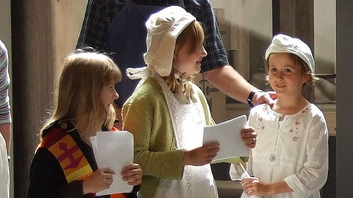 Fotoaufnahme einer Szene aus dem Theaterstück zum Thema Gründung und Leben eines mittelalterlichen Spitals. Im Zentrum stehen drei kostümierte Mädchen mit Textblättern in der Hand. Dahinter sind zwei Erwachsene, die das Schauspiel beobachten.