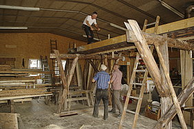 Zimmermänner sägen mit einer Schrotsäge Balken zu Brettern. Dabei stehen zwei Zimmerer unterhalb eines Gerüsts, auf dem der Balken aufliegt, ein Zimmerer steht oben.