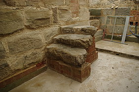 Fotoaufnahme der drei Sandsteinstufen, die vom Flur in die tieferliegende Schürkammer führen. Die Stufen befinden sich an der Mauer aus Sandsteinquadern und wurden mit Ziegelsteinen untermauert. Im Hintergrund ist eine weitere Mauer erkennbar, aus der zwei Rohre führen. 