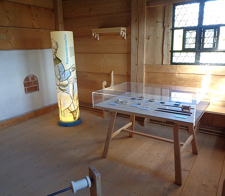 ein Tisch mit Exponaten, durch Glas geschützt sowie ein stehender Zylinder mit Ausstellungstext