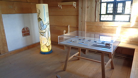 ein Tisch mit Exponaten, durch Glas geschützt sowie ein stehender Zylinder mit Ausstellungstext
