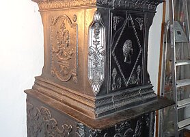 Der Ofen ist schwarz. Er besteht aus zwei aufeinanderstehenden Kästen. Oben ist ein geschwungen gestalteter Abschluss. Der Ofen steht auf zwei Füßen. Sonst ist er an der Wand angebaut.