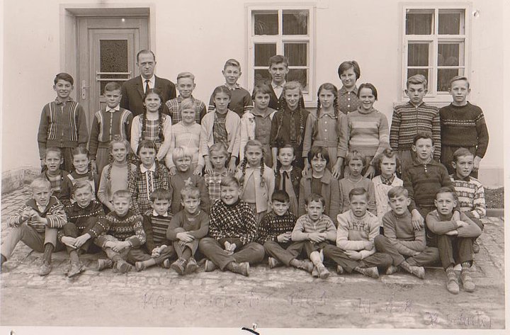 Historische schwarz-weiße Fotoaufnahme einer Schulklasse vor einem Haus. Die Klasse hat sich zum Klassenfoto in vier Reihen aufgestellt, die vorderen beiden Reihen sitzen bzw. knien. Der Lehrer steht links in der letzten Reihe. 