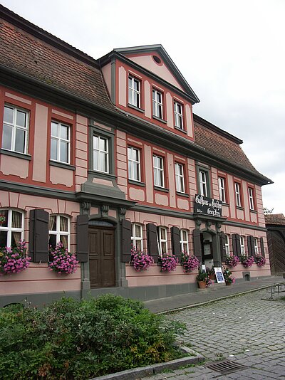 FFM_Gasthof zum Hirschen Ein repräsentatives, zweigeschossiges Gasthaus, rosa gestrichener Putz mit roten Fenstereinfassungen. Im Erdgeschoss zieren die Fensterkästen lila Blumen