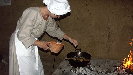Fotoaufnahme einer Frau in mittelalterlichem Gewand mit Schürze und Haube. Vor ihr steht ein Metalltopf über heißen Kohlen, in dem sie etwas zubereitet. Daneben brennt ein Feuer. Auf dem Herd steht außerdem eine kleine Kanne. 