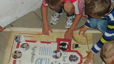 Fotoaufnahme der Schatzsuche in der Spitalkirche in Bad Windsheim. Die aufgerollte Schatzkatze ist auf den Holzdielen des Bodens ausgebreitet. Die Kinder sitzen bzw. stehen um die Karte und studieren sie. 
