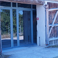 Fotoaufnahme des Eingangs vom Archäologiemuseum, welches sich in der Schafscheune aus Virnsberg befindet. Das zweiflüglige Holztor ist nach außen geöffnet, die beiden modernen Glastüren dahinter sind geschlossen. Neben der rechten Tür an der Wand befindet sich ein Handfeuermelder.