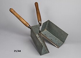 Die Schere hat Holzgriffe. An den Schneiden sind Blechkästen angebracht. Der Blechkasten schließt sich, wenn die Schere geschlossen wird.