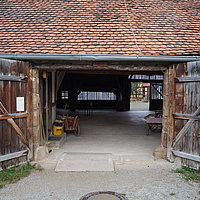 Fotoaufnahme des hinteren Eingangs vom Alten Bauhof aus Bad Windsheim. Das zweiflügelige Holztor ist nach außen geöffnet. An der rechten Seite hängt ein „Rauchen verboten“-Schild. Das Dach nimmt das obere Drittel des Bildes ein und ist mit Sechseck-Flachziegeln gedeckt. 