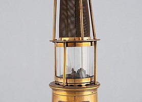 Die Lampe ist aus Messing. Sie ist goldfarben. In der Mitte ist ein Glasgehäuse. Darüber ist ein rundes Metallgitter. Die Lampe hat einen Griff.