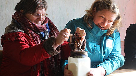 Fotoaufnahme von zwei Frauen beim Buttern, als Teil der aktiven Führung für Erwachsene zum Thema Milch. Auf einem Tisch steht ein Butterglas, auf der Bank dahinter sitzen zwei lachende Frauen. Die Frau rechts hält das Glas fest, während die Frau links an der Kurbel dreht.