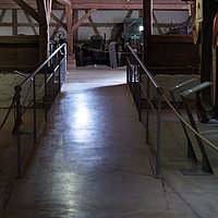 Fotoaufnahme der langgezogenen Rampe mit Geländer im Innenraum von der Schafscheuer aus Weiltingen. Hinter der Rampe sind die Fachwerk-Fassade, sowie der offene Dachstuhl erkennbar. In dem Raum werden verschiedene Maschinen ausgestellt.  
