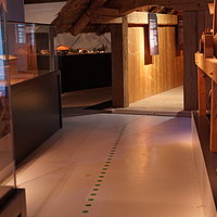 Fotoaufnahme der Ausstellung des Archäologiemuseums, welches sich in der Schafscheune aus Virnsberg befindet. Ein Leitsystem auf dem Boden führt durch die Ausstellung, links und rechts davon stehen Vitrinen mit Objekten. 