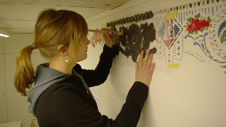 Fotoaufnahme der Schablonenmalerei im Bauernhaus aus Mailheim. Eine junge Frau steht vor einer Wand, an die mit Klebeband eine Schablone geklebt wurde. Mit entsprechender Stupftechnik bringt sie Muster in verschiedenen Farben an der Wand auf. 