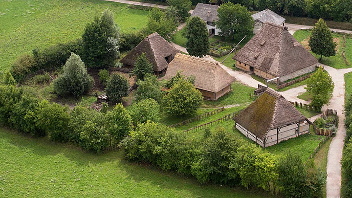 Fotoaufnahme eines Luftbildes der Baugruppe Mittealter. Die Gruppe von Häusern auf einer Wiese ist von einer Hecke aus Bäumen umgeben und über Wege zugänglich. Umliegend ist die Wiese erkennbar. 