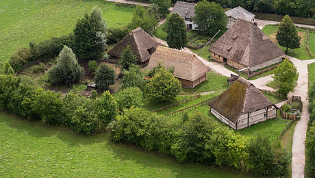Fotoaufnahme eines Luftbildes der Baugruppe Mittealter. Die Gruppe von Häusern auf einer Wiese ist von einer Hecke aus Bäumen umgeben und über Wege zugänglich. Umliegend ist die Wiese erkennbar. 