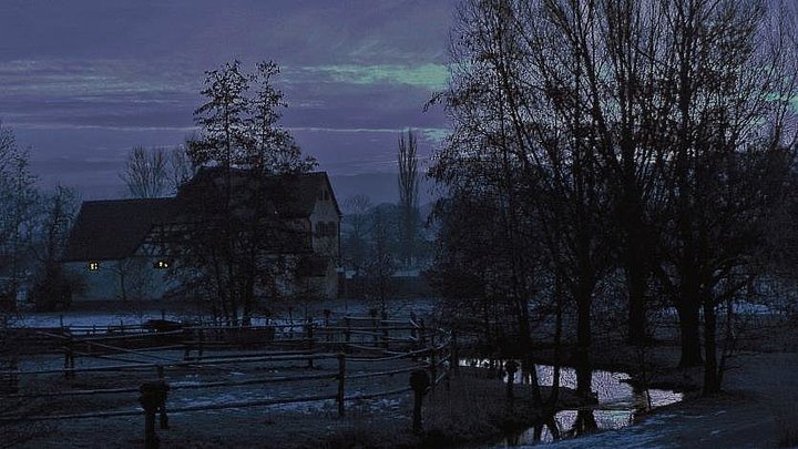 Fotoaufnahme vom Freilandmuseum bei Nacht. Im Vordergrund befindet sich das schneebedeckte Ufer eines kleinen Baches. Links dahinter ist eine eingezäunte Weide, rechts stehen kahle Bäume. Im Hintergrund ist das Köblerhaus aus Schwimbach erkennbar. 