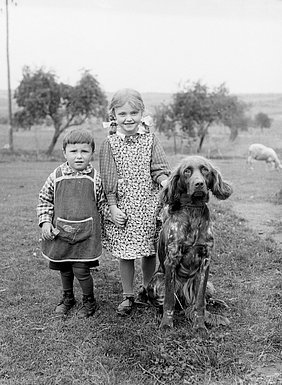 Ein Mädchen und ein kleinerer Junge stehen neben einem Hund. Sie sind auf einer Wiese. Beide Kinder tragen Schürzen. Die Schürze vom Mädchen hat ein Blumenmuster.