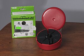 Der Telefonkabelaaufroller ist eine runde Dose. Sie ist rot und aus Plastik. Daneben liegt die Verpackung. Sie ist grün. Sie zeigt eine Frau beim Telefonieren.