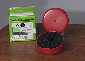 Der Telefonkabelaaufroller ist eine runde Dose. Sie ist rot und aus Plastik. Daneben liegt die Verpackung. Sie ist grün. Sie zeigt eine Frau beim Telefonieren.