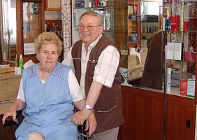 In der Mitte steht der Friseur Josef Stingl. Er trägt ein Hemd und eine braune Weste. Links von ihm sitzt seine Ehefrau Franziska Stingl. Sie trägt einen hellblauen Kittel. Im Hintergrund sieht man Möbel des Friseurgeschäfts. Es sind Schränke, Vitrinen und Spiegel.