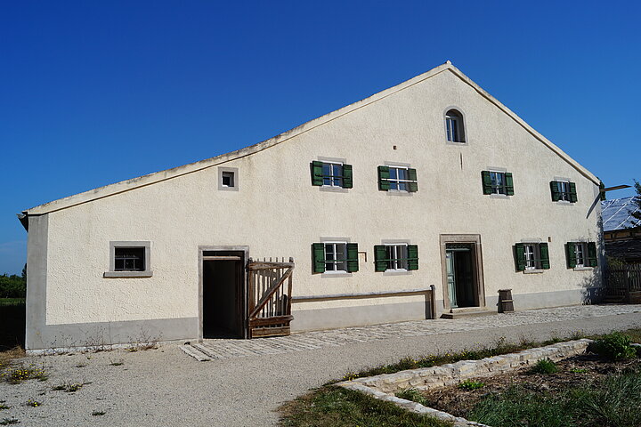 Fotoaufnahme des Bauernhauses aus Reichersdorf am aktuellen Standort. Das breitgelagerte eingeschossige Jurahaus hat zwei Eingänge an der Giebelseite. Links ist die Stalltür, rechts die Haustür aus Holz. Die Fenster auf der Wohnseite haben geöffnete Läden. 