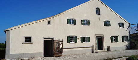 Fotoaufnahme des Bauernhauses aus Reichersdorf am aktuellen Standort. Das breitgelagerte eingeschossige Jurahaus hat zwei Eingänge an der Giebelseite. Links ist die Stalltür, rechts die Haustür aus Holz. Die Fenster auf der Wohnseite haben geöffnete Läden. 