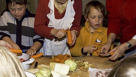 Fotoaufnahme der Aktionstage „Ernährung“. Um einen Holztisch steht eine Gruppe Kinder und schneidet Gemüse. Auf dem Tisch liegen Karotten, Kartoffeln, Zwiebeln, Kohlrabi und Sellerie. Zwei Frauen helfen den Kindern beim Karottenschälen. 