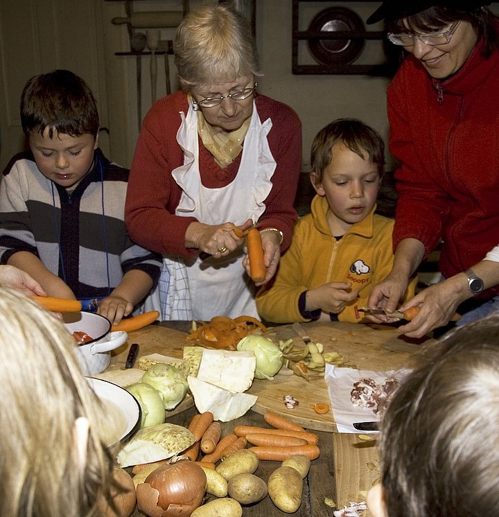 Fotoaufnahme der Aktionstage „Ernährung“. Um einen Holztisch steht eine Gruppe Kinder und schneidet Gemüse. Auf dem Tisch liegen Karotten, Kartoffeln, Zwiebeln, Kohlrabi und Sellerie. Zwei Frauen helfen den Kindern beim Karottenschälen. 