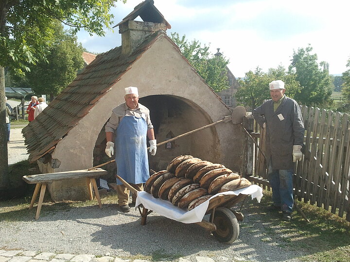 Fotoaufnahme des Backofens aus Haidling während des Backofenfestes. Vor dem kleinen Häuschen stehen zwei Männer mit einer Schubkarre aus Holz. Diese ist vollbeladen mit frischgebackenen Brotlaiben. Im Hintergrund sind mehrere Besucher, Bäume und Fachwerkhäuser erkennbar. 