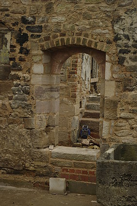 Fotoaufnahme der Türöffnung zwischen Flur und Badstube. Ein gemauerter Korbbogen ist in das Steinmauerwerk eingebettet. Rechts neben der Tür steht eine steinerne Wanne. Im Raum dahinter ist eine Treppe aus Sandsteinquadern erkennbar, auf der eine Kabeltrommel steht. 