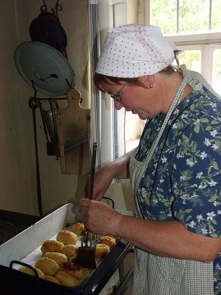 Fotoaufnahme vom Kochen und Backen. Eine Frau mit Schürze und Kopftuch steht in einer Küche. Vor ihr steht eine Auflaufform, in der sich gebackene Teigbällchen befinden. Die Frau dreht diese gerade mit einem Pfannenwender um. 