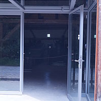 Fotoaufnahme vom verglasten Eingangsbereich der Schafscheuer aus Weiltingen am aktuellen Standort. Im Gebäude sind der Sichtdachstuhl, sowie einige Maschinen erkennbar. Der Bereich der Exponate wird mit einem Seil abgegrenzt. 