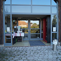 Fotoaufnahme des Eingangsbereichs zur Ausstellungsscheune bzw. dem Stadel aus Betzmannsdorf. Das zweiflüglige Holztor ist nach außen geöffnet, dahinter liegt der verglaste Eingangsbereich mit geschlossener Tür. Auf dem grob gepflasterten Hof davor steht ein Aschenbecher.