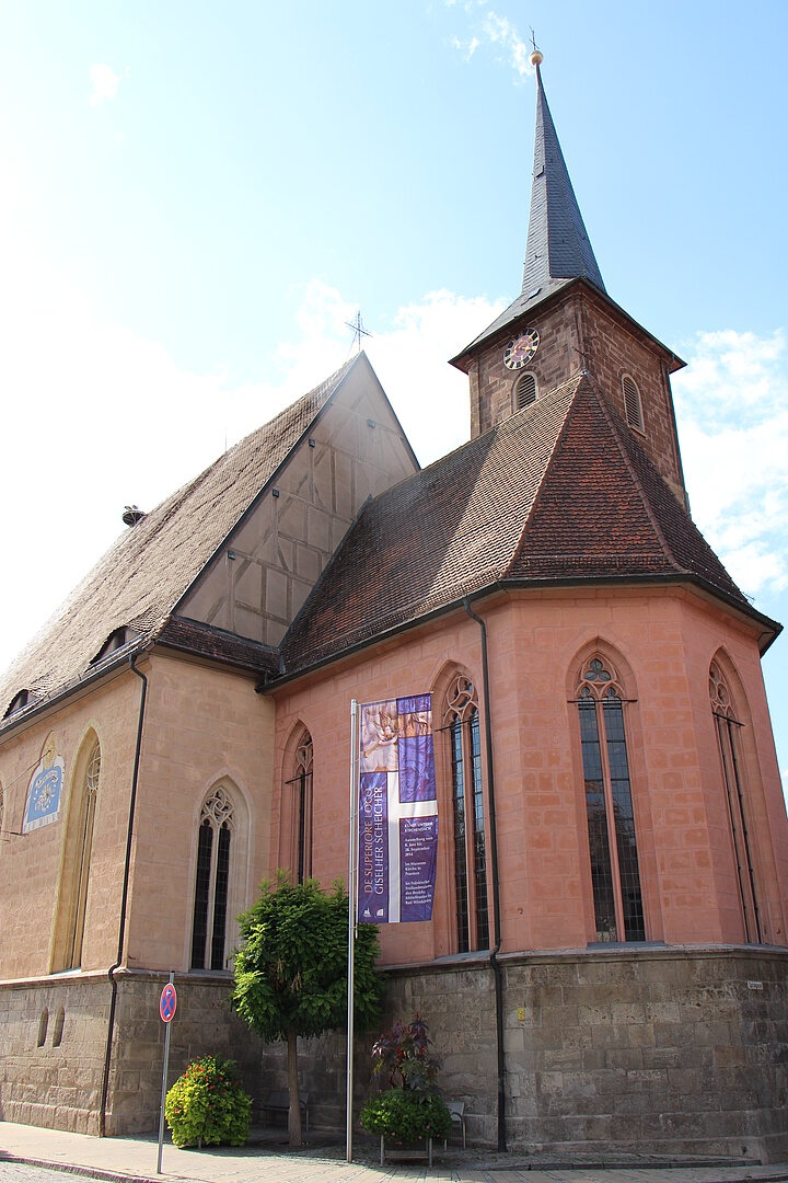 Fotoaufnahme der Spitalkirche in Bad Windsheim von außen. Die Kirche mit schmalen hohen Fenstern, dem Fachwerkgiebel und spitzem Kirchturm wird von der Sonne beschienen. Davor weht eine Fahne der Ausstellung „Kunst unterm Kirchendach“, daneben stehen zwei Büsche und ein Baum.