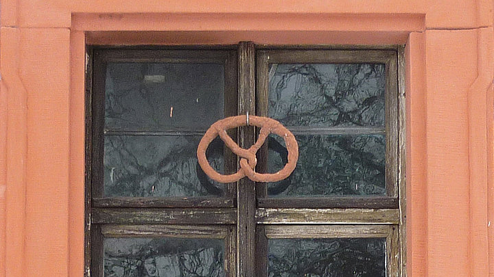 Fotoaufnahme des Fensters der Backstube im Amtshaus aus Obernbreit. Das Fenster ist in einem steinernen Rahmen eingebettet. Am hölzernen Fensterkreuz ist eine Brezel angebracht. 