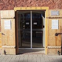 Fotoaufnahm der geöffneten Eingangstür zur Museumskasse im Korbhaus aus Knittelsbach am aktuellen Standort. In den Türrahmen wurden die Jahreszahl „2006“, sowie das Fränkische Wappen eingeprägt. Hinter der Holztür befindet sich eine automatische Schiebetür. 