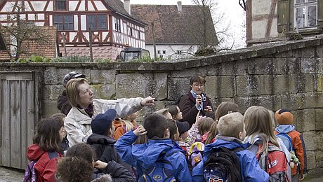 Fotoaufnahme einer Gruppe Schulkinder, die vor dem Bauernhaus aus Herrnberchtheim steht. Hinter der Hofmauer ist neben kahlen Bäumen unter anderen das Amtshaus aus Obernbreit erkennbar. Die Führerin zeigt auf etwas außerhalb des Bildes. 