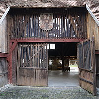 Fotoaufnahm des vorderen Eingangs zum  Alten Bauhof aus Bad Windsheim. Die rechte Seite des zweiflügligen Holztors ist geöffnet. Darüber hängt ein Wappen mit einem Adler und einem „W“ in der Mitte. Im Inneren sind einige Objekte erkennbar. 