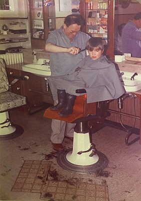 Das Bild zeigt einen Friseursalon. In der Mitte ist ein Friseurstuhl. Auf dem Stuhl sitzt ein kleiner Junge. Er hat einen grauen Kittel an. Der Friseur schneidet dem Jungen die Haare. Auch der Friseur trägt einen grauen Kittel. Im Hintergrund sind Waschbecken und Spiegel.