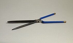 Das Onduliereisen sieht aus wie eine Schere. Es hat blaue Griffe mit goldfarbenen Enden. Das eine Eisen ist ein runder Stab. Das andere Eisen ist eine hohle Form. In sie passt der runde Stab hinein.