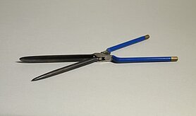 Das Onduliereisen sieht aus wie eine Schere. Es hat blaue Griffe mit goldfarbenen Enden. Das eine Eisen ist ein runder Stab. Das andere Eisen ist eine hohle Form. In sie passt der runde Stab hinein.