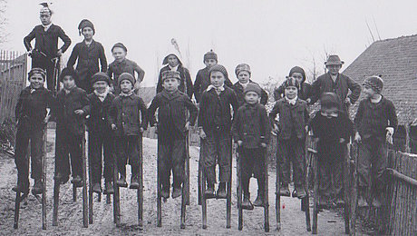 Historische schwarz-weiße Fotoaufnahme einer 20-köpfigen Kindergruppe auf hölzernen Stelzen. Sie stehen in zwei Reihen hintereinander auf einem Feldweg in einem Dorf. Alle Kinder tragen Jacken und Mützen. Links im Hintergrund sind kahle Bäume erkennbar, rechts ein Haus. 