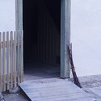 Fotoaufnahme des Haupteingangs vom Bauernhaus aus Kleinrinderfeld am aktuellen Standort. Die Tür ist nach innen geöffnet, vor der Schwelle liegt eine Rampe aus Holz. Links neben der Tür befindet sich ein hölzernes Gatter, rechts ein Reisigbesen und ein kleines Fenster. 