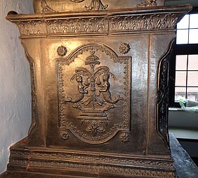 An der Seite des Ofens ist ein Bild. Es zeigt zwei Wasserfrauen. Sie stehen um einen Springbrunnen herum.