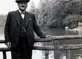 Schwarz-Weiß Fotografie von einem Mann im Anzug mit Hut, an einem Geländer lehnend, vor einem Gewässer. 